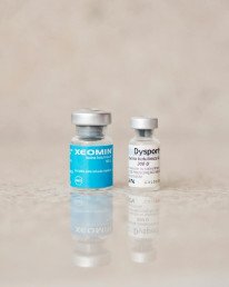 Close-up of Xeomin and Dysport bottles reflecting on a surface, shot indoors.